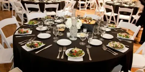 A round banquet table set with black linens, white plates, silverware, and a salad dish in a large event venue.