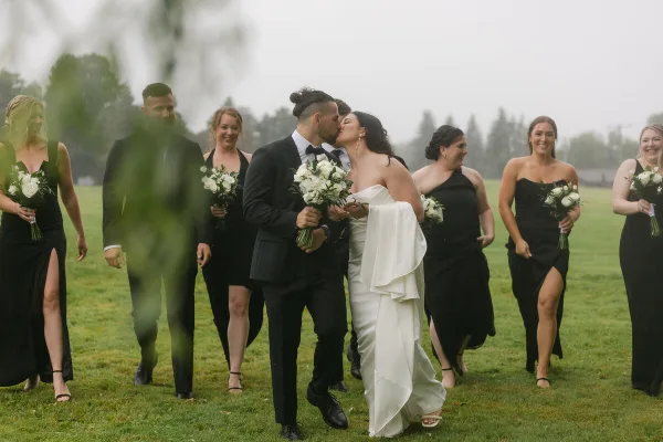 A bride and groom kiss in a grassy field, surrounded by their wedding party.