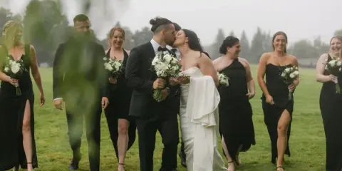 A bride and groom kiss in a grassy field, surrounded by their wedding party.