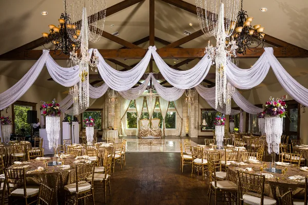 Wedding reception hall decorated with white drapery, chandeliers, floral arrangements, and set dining tables.