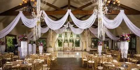 Wedding reception hall decorated with white drapery, chandeliers, floral arrangements, and set dining tables.