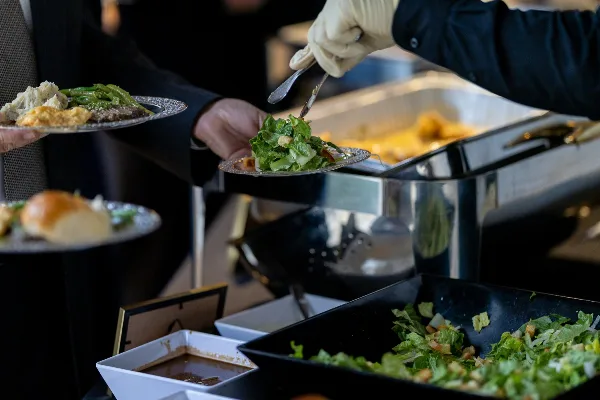 A server wearing a white glove uses a serving utensil to place a salad onto a plate held by a guest at a catering event.