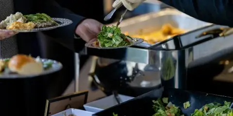 A server wearing a white glove uses a serving utensil to place a salad onto a plate held by a guest at a catering event.