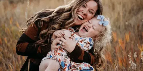 A smiling woman holding a laughing young child in a field of tall dry grass.