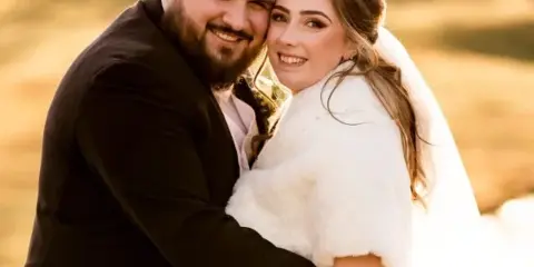 A professional wedding photograph of a smiling bride and groom embracing outdoors.