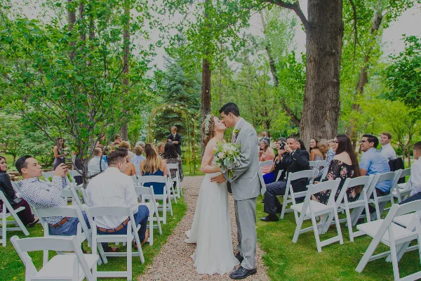 A bride and groom share a kiss during an outdoor wedding ceremony in a garden setting with seated guests.