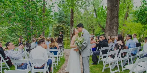 A bride and groom share a kiss during an outdoor wedding ceremony in a garden setting with seated guests.
