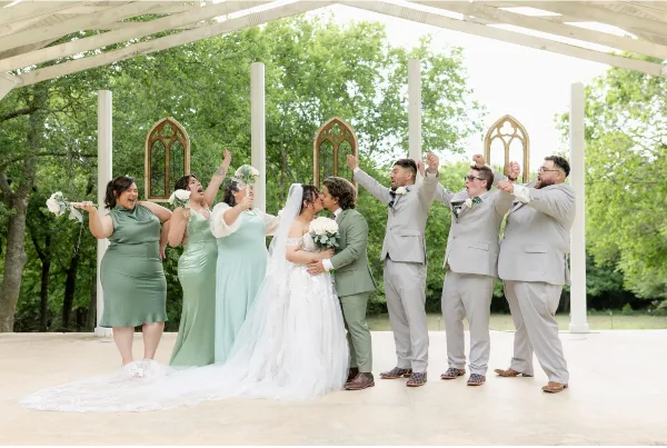 A bride and groom kissing surrounded by their wedding party under a white wooden pergola structure.