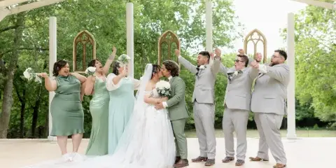 A bride and groom kissing surrounded by their wedding party under a white wooden pergola structure.