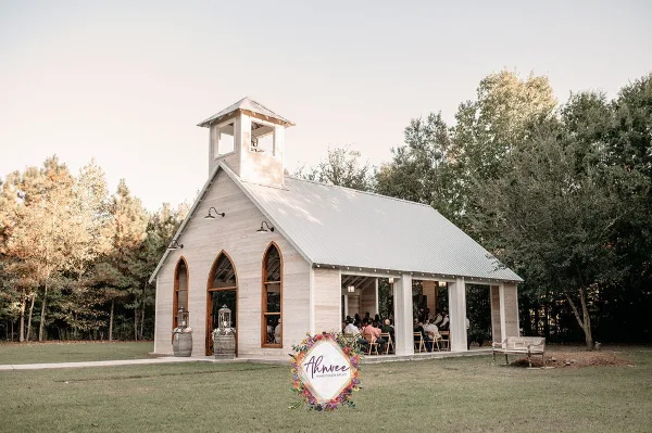 A white open-air wedding chapel with a bell tower and arched stained-glass windows surrounded by trees and a grassy lawn.