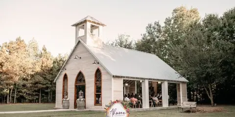 A white open-air wedding chapel with a bell tower and arched stained-glass windows surrounded by trees and a grassy lawn.