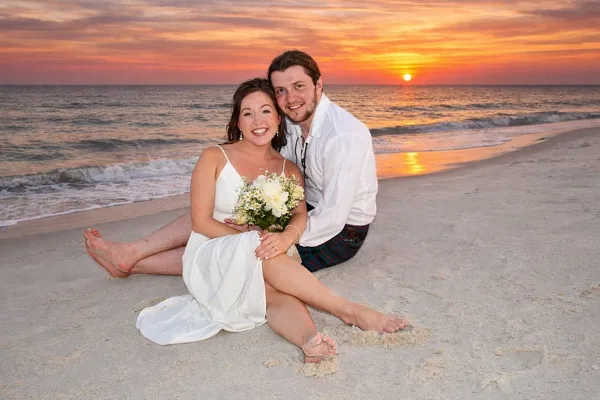 A bride and groom sitting on a sandy beach holding a bouquet of white flowers with a sunset over the ocean in the background.