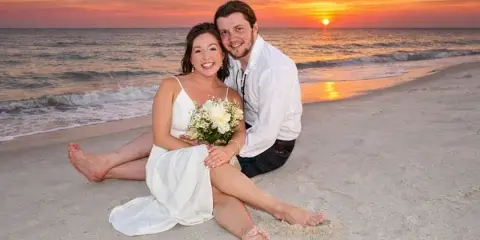 A bride and groom sitting on a sandy beach holding a bouquet of white flowers with a sunset over the ocean in the background.