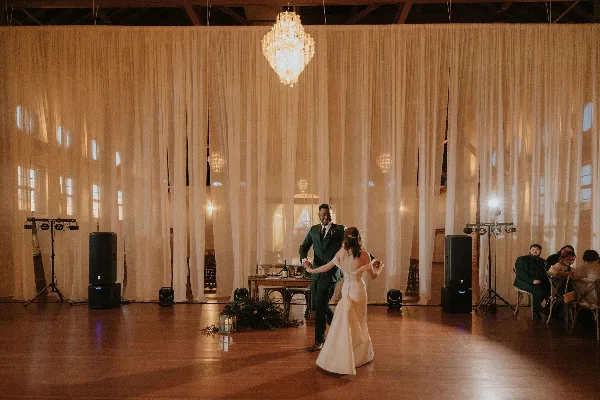 A bride and groom dancing at a wedding reception in a venue with white drapery and a chandelier.
