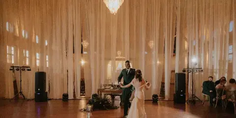 A bride and groom dancing at a wedding reception in a venue with white drapery and a chandelier.