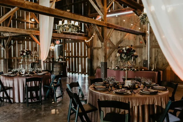 Interior view of a rustic barn wedding venue featuring exposed wooden beams, chandeliers, and round tables set with tablecloths and floral centerpieces.