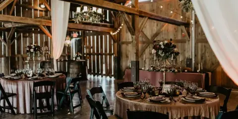 Interior view of a rustic barn wedding venue featuring exposed wooden beams, chandeliers, and round tables set with tablecloths and floral centerpieces.