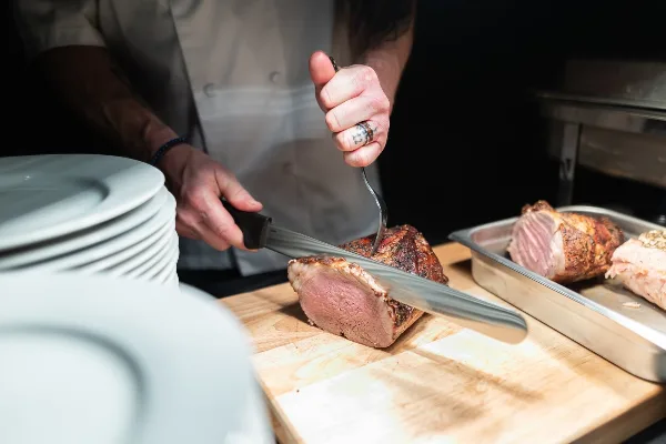 Chef in white uniform carving a roasted meat on a wooden board with stacked white plates nearby