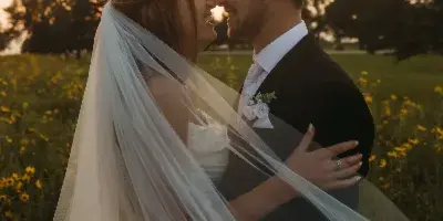 A bride and groom sharing a moment in a field of yellow flowers at sunset, with the bride's veil blowing in the wind.
