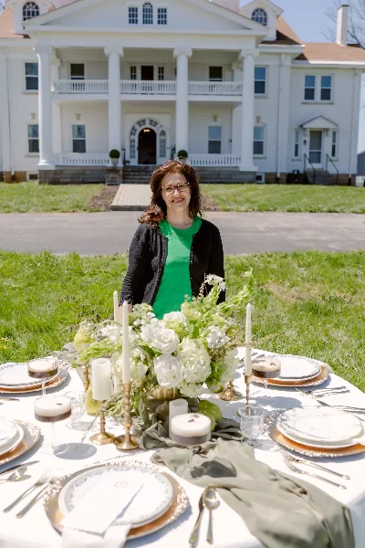 A woman stands behind a decorated wedding table with white flowers, candles, and fine china in front of a white building.