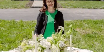 A woman stands behind a decorated wedding table with white flowers, candles, and fine china in front of a white building.
