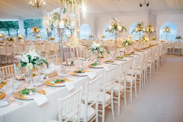 Interior of a white event tent featuring long banquet tables with white linens and rows of white Chiavari chairs.