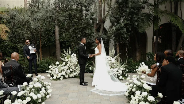 A bride and groom standing at the altar during an outdoor wedding ceremony, surrounded by white floral arrangements and guests.