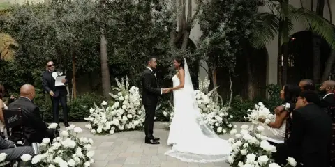 A bride and groom standing at the altar during an outdoor wedding ceremony, surrounded by white floral arrangements and guests.