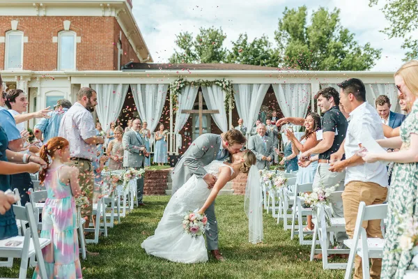 A bride and groom share a kiss at the end of their wedding aisle while guests throw flower petals.