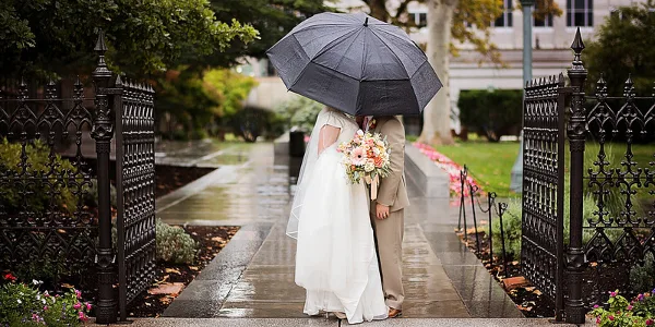 A bride and groom standing under a black umbrella on a wet stone pathway, with the bride holding a bouquet.