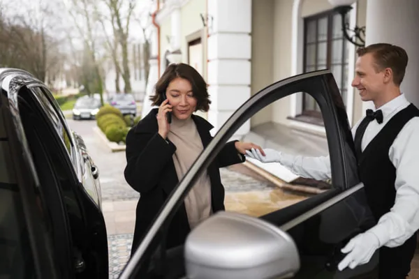 A professional chauffeur in a white shirt and bow tie opens the door of a black luxury sedan for a passenger.