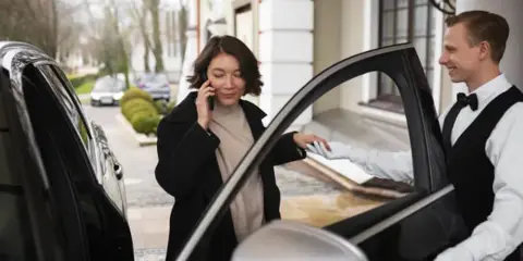 A professional chauffeur in a white shirt and bow tie opens the door of a black luxury sedan for a passenger.