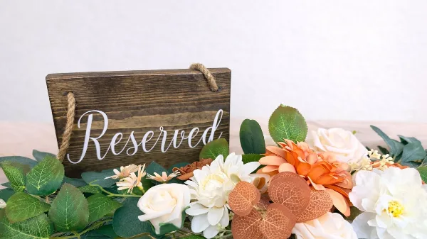 A rustic wooden 'Reserved' sign surrounded by a lush arrangement of white and orange flowers and greenery, representing event decor styling.