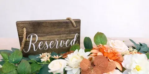 A rustic wooden 'Reserved' sign surrounded by a lush arrangement of white and orange flowers and greenery, representing event decor styling.