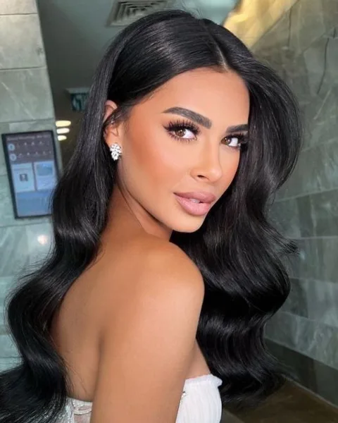 A woman with long, dark wavy hair and professional makeup looking over her shoulder.