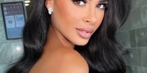 A woman with long, dark wavy hair and professional makeup looking over her shoulder.