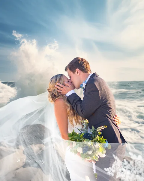 A bride and groom kissing on a beach with waves crashing in the background.