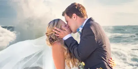 A bride and groom kissing on a beach with waves crashing in the background.