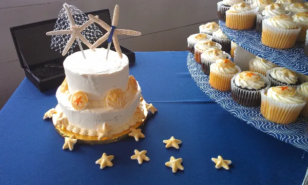 A two-tiered white wedding cake decorated with starfish and seashells sits next to a tiered stand of frosted cupcakes on a blue table.