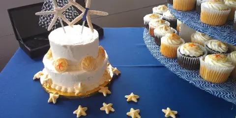 A two-tiered white wedding cake decorated with starfish and seashells sits next to a tiered stand of frosted cupcakes on a blue table.