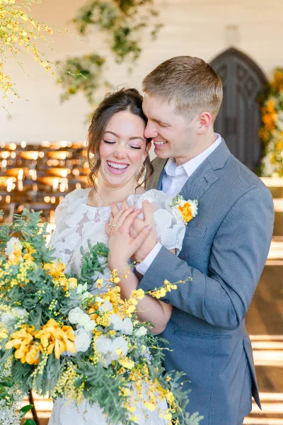 A bride and groom smiling and holding hands during a wedding ceremony, with the bride holding a bouquet of yellow and white flowers.