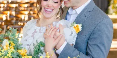 A bride and groom smiling and holding hands during a wedding ceremony, with the bride holding a bouquet of yellow and white flowers.