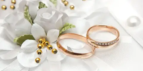 Rose gold wedding rings and a white floral boutonniere resting on white fabric.
