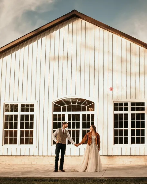 A bride and groom holding hands in front of a white barn-style wedding venue with large arched windows.