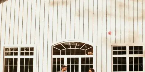 A bride and groom holding hands in front of a white barn-style wedding venue with large arched windows.
