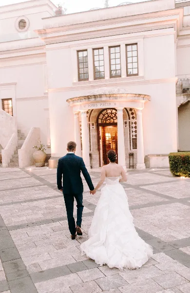 A bride and groom walking hand-in-hand across a stone courtyard toward a white building entrance.