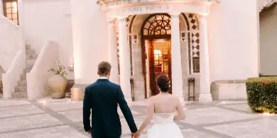 A bride and groom walking hand-in-hand across a stone courtyard toward a white building entrance.