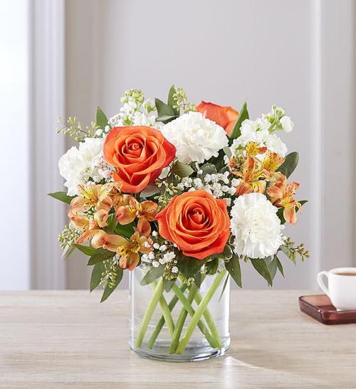 A floral arrangement featuring orange roses, white carnations, and alstroemeria in a clear glass vase.