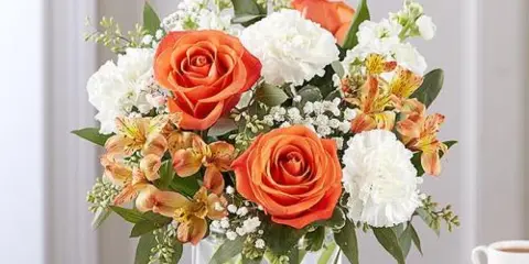 A floral arrangement featuring orange roses, white carnations, and alstroemeria in a clear glass vase.