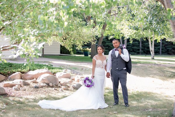 A bride and groom standing outdoors in a garden setting, holding hands and posing for a wedding photograph.